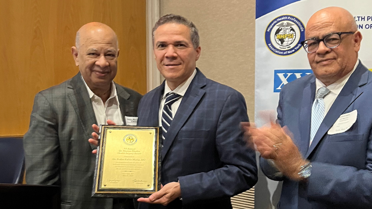 Dr. Pedro P. Maria, DO, Attending Physician and Associate Professor of Urology, dressed in a blue suit and striped tie, holds up an award plaque. Smiling colleagues stand to his right and left.
