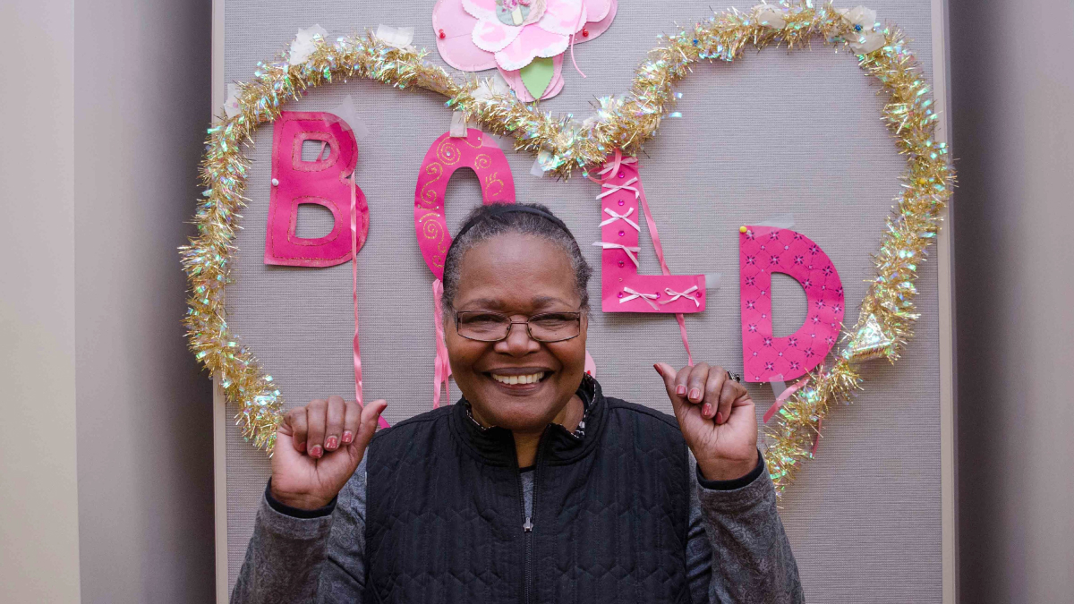 A BOLD cancer survivor stands in front of a decorated celebration wall, holding her hands up victoriously.