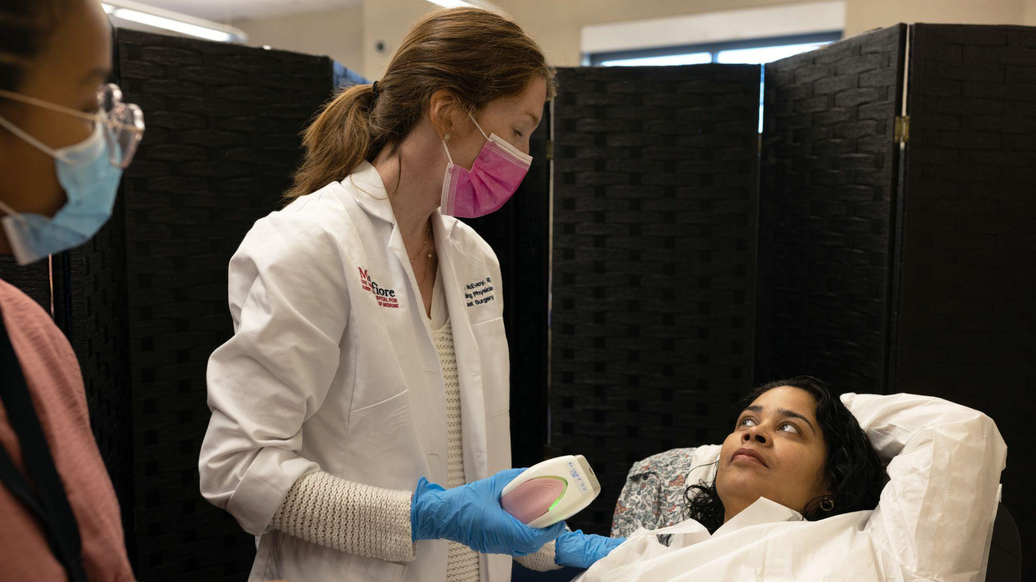 Two medical professionals consult with a patient in a private screened area.  The doctor holds the iBreastExam device.