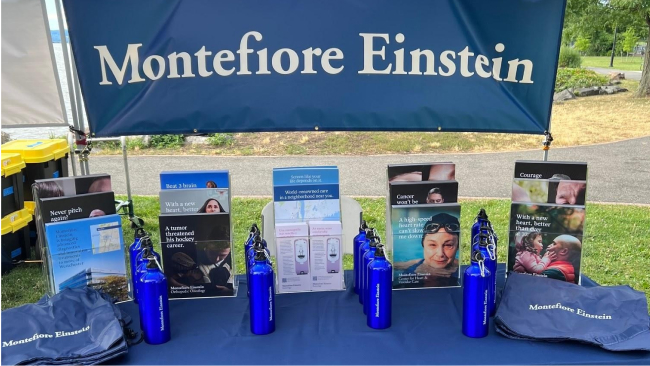 A table draped in a blue tablecloth, displaying informational flyers, promotional bags, and water bottles all sporting the Montefiore Einstein logo. A banner behind the display reads Montefiore Einstein in white text on a blue background.