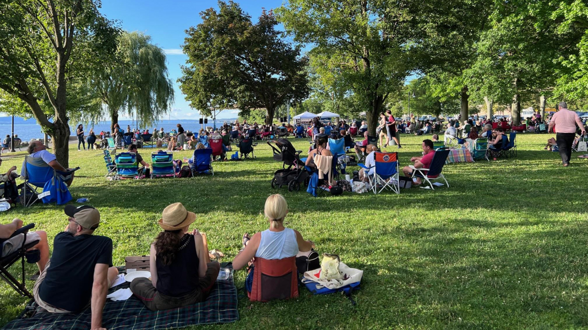Groups of people sit on sun dappled green grass, surrounded by shady trees, to watch the jazz festival in the distance.