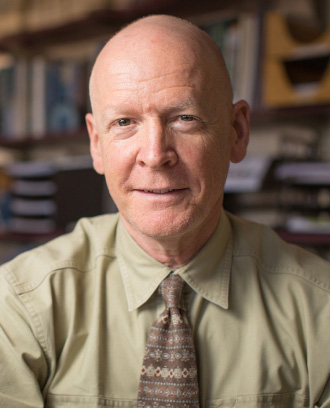 Peter A. Selwyn, MD, MPH, wearing a tan shirt and brown tie. Shelves full of books are behind him.