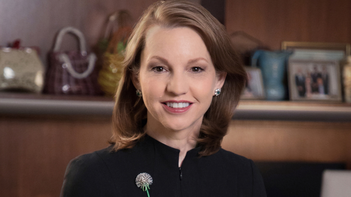 Susan Fox, President and CEO. She smiles toward the camera, wearing a black shirt and small silver brooch.  Behind her is a wooden mantle with books and ceramics.
