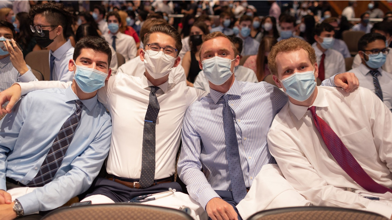 Four masked med students in button down shirts and ties lean together for a picture. 