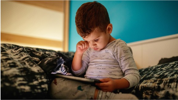 A toddler rubs their eye as they view a tablet in a dim room.