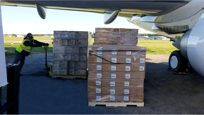 Two pallets piled high with supplies are stacked in the shade of the jet's wing.