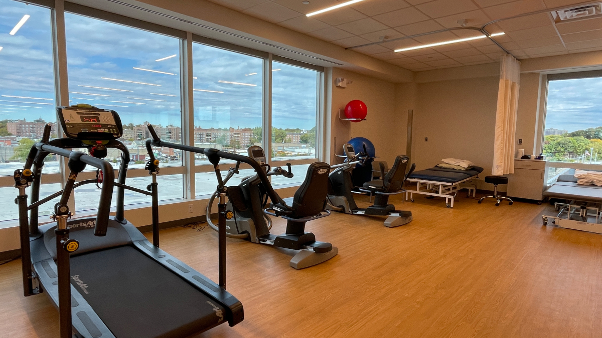 New rehabilitation and exercise equipment along the walls of a bright room with floor to ceiling windows and a hardwood floor.