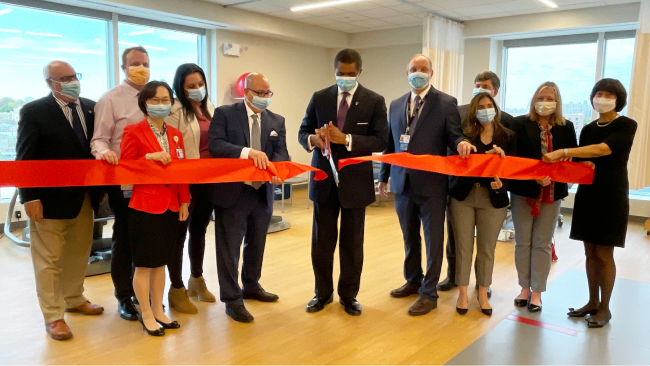 A team of doctors and admin behind a big red ribbon. Dr. Ozuah cuts the ribbon with large scissors. 