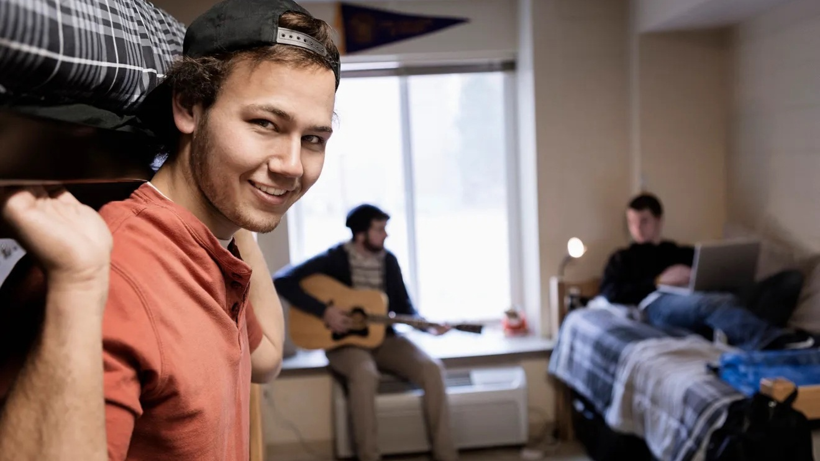 A young man in a light red shirt smiles toward the camera, leaning out from under a bunk bed. Behind him, two other young men play a guitar and work on a computer in this dorm room setting.