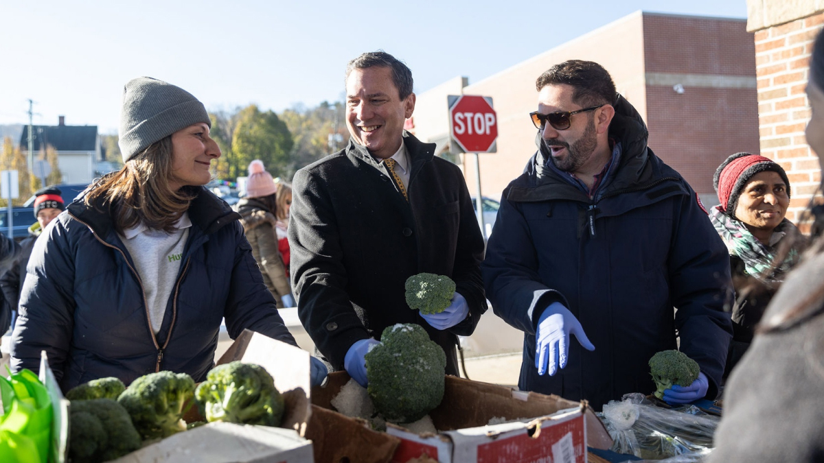 Three people dressed in warm winter clothes smile and talk in front of a set of boxes of greens and brocolli.