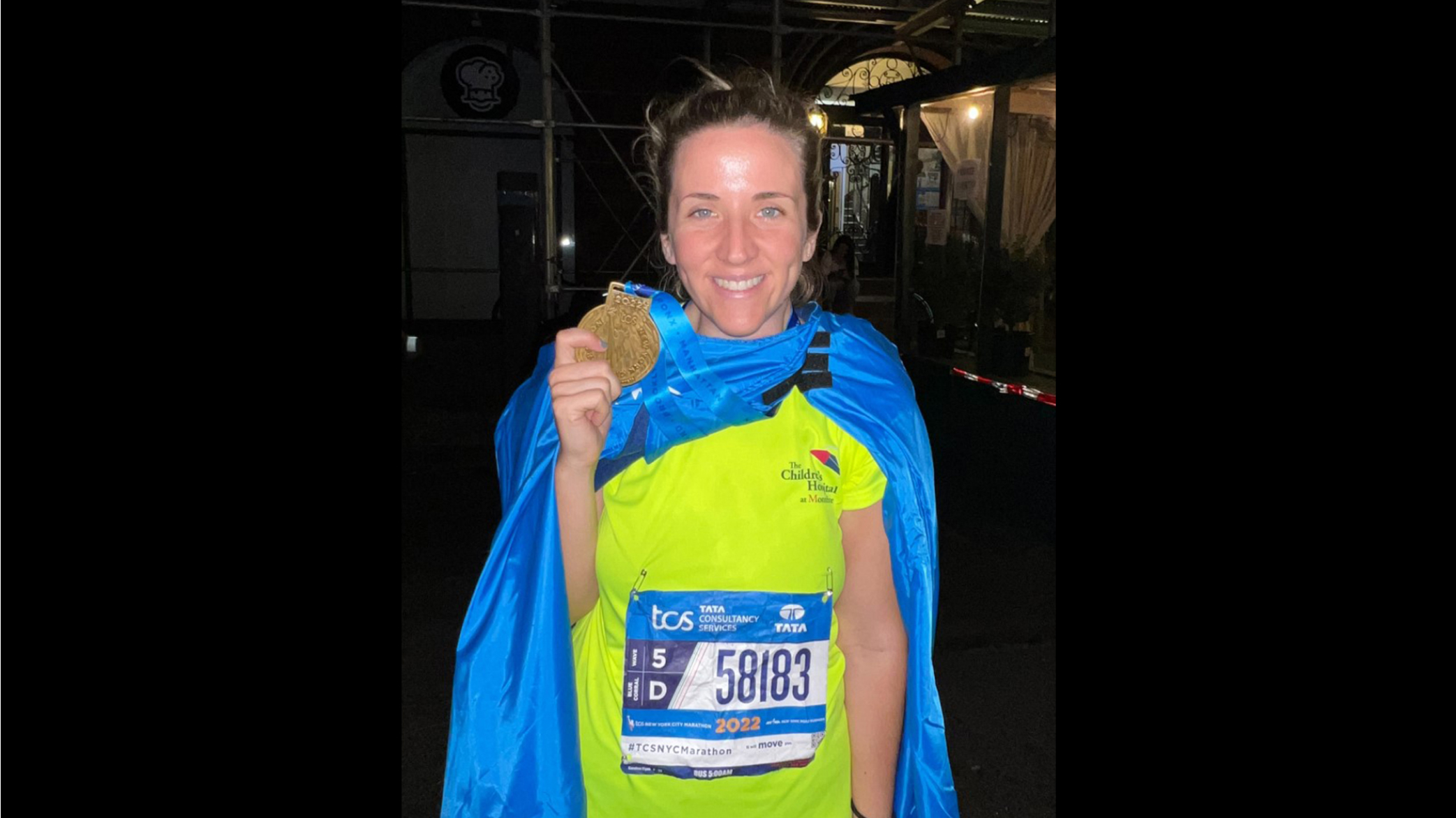 A runner in a neon yellow shirt and blue cape/blanket holds up their golden medal with a proud smile.