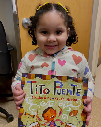 An adorable child smiles while holding up a book about Tito Puente.