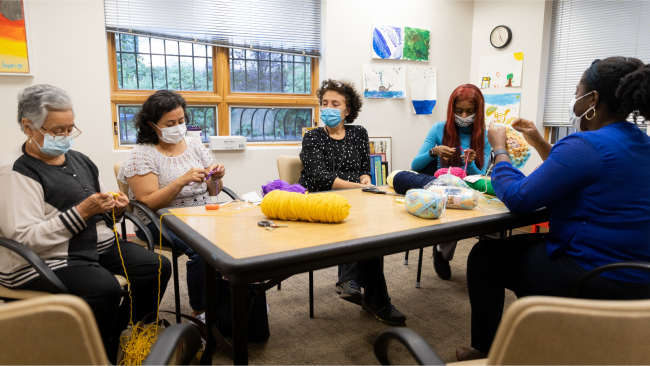 A group of patients and doctors sit around a table covered in colorful yarn, working on crochet projects.  