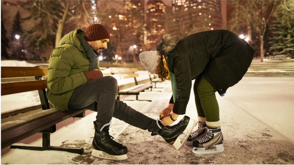 Two ice skaters lace up their skates in a wintery scene.