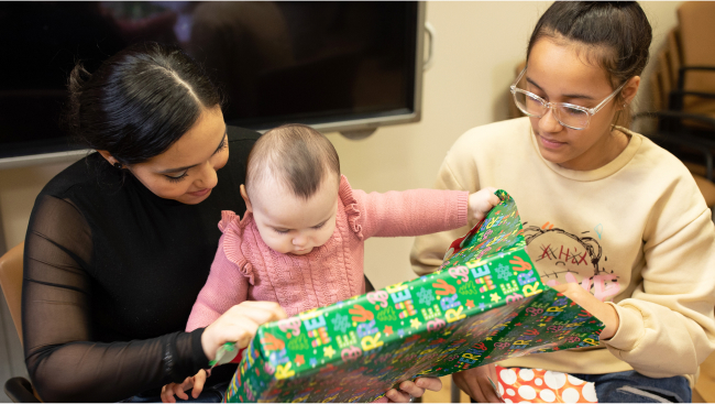 Two women help a baby open present wrapped in green and red paper.