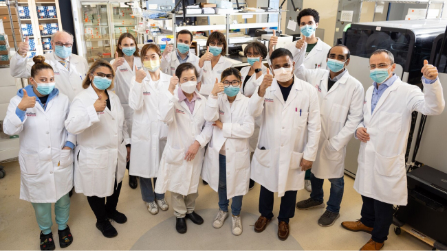A group of scientists in masks and white lab coats pose for the camera.