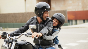 A parent and child, wearing helmets, ride a motorcycle together.