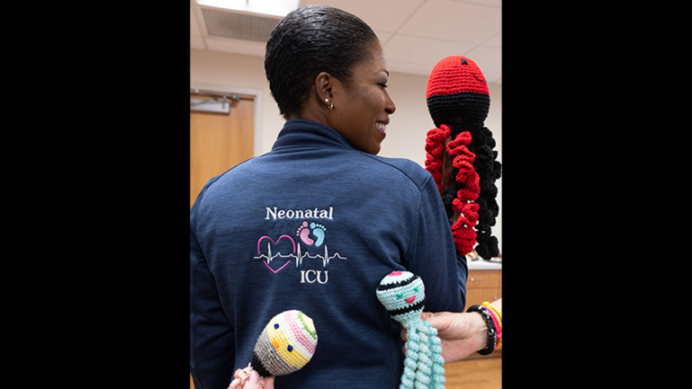 A staffer wears an embroidered jean shirt featuring the NICU logo. From off camera, a hand holding a crochet baby toy.