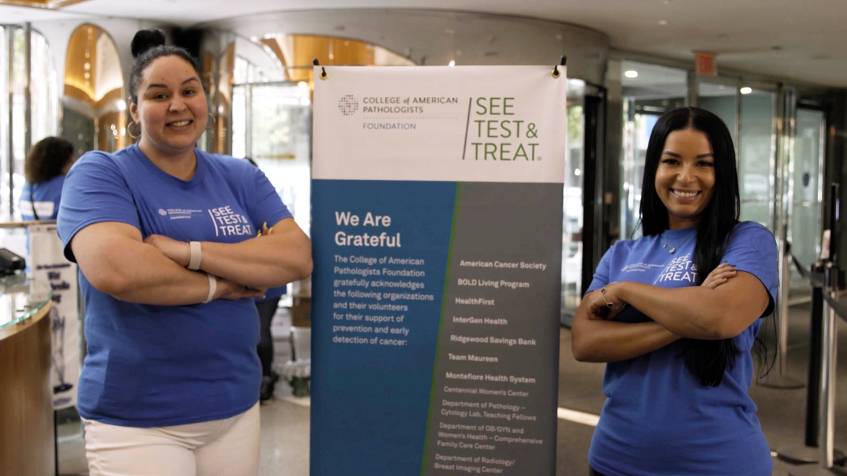Two medical professionals in matching t-shirts stand to either side of the See, Test & Treat event sign.