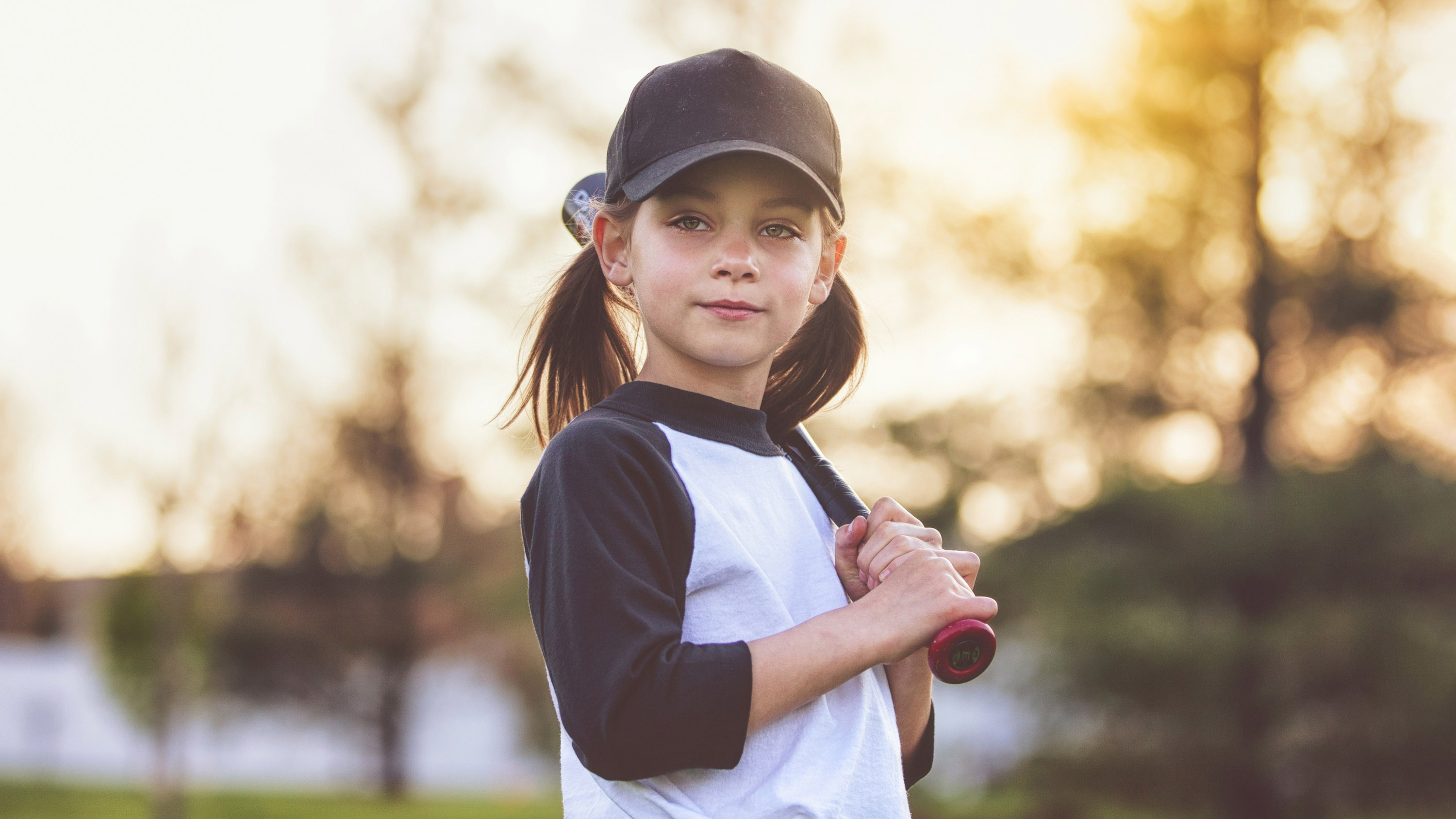 A girl in a baseball hat and little league shirt holds a bat on her shoulder.