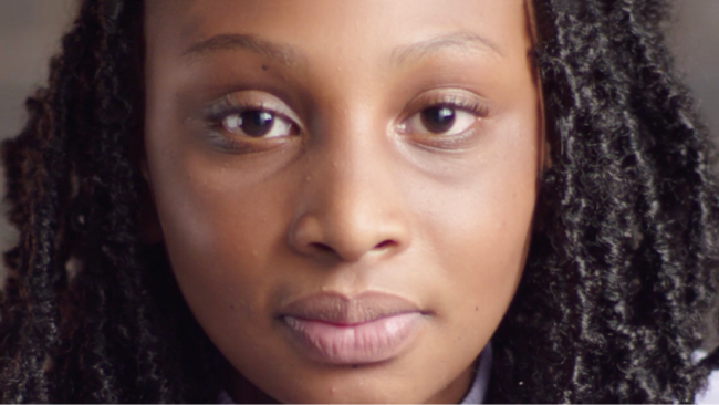A young Black woman looks directly into the camera.  Her expression is resolute.