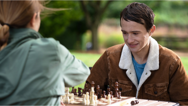 Two young adults play chess outdoors on a brisk day.