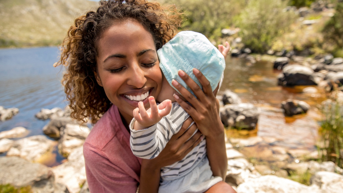 A woman hugs a baby close and smiles in a sunny outdoor scene with a lake in the background.