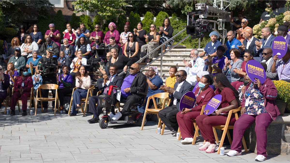 A crowd of about two dozen people sits to either side of Dr. Gresham, who is seated in the front of a sunny outdoor courtyard.