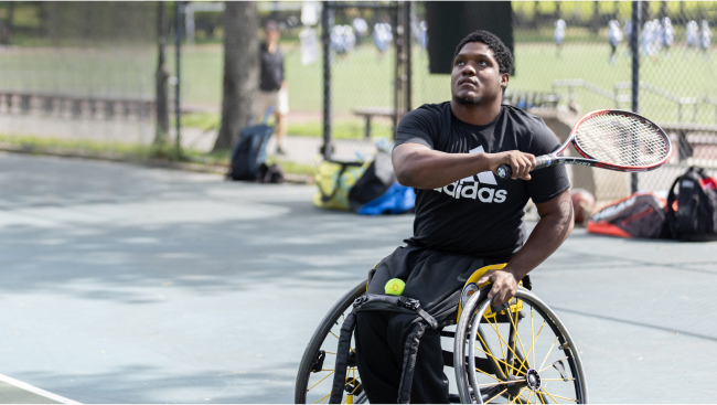 An athlete in a wheelchair prepares to backhand a tennis ball on the court.