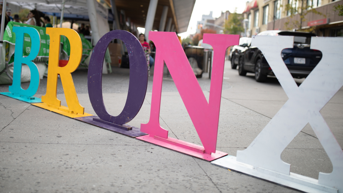 A sculptural metal sign stands on the sidewalk. It's a series of letters - B in aqua, R in yellow, 0 in navy, N in pink, and X in white, spelling BRONX. 