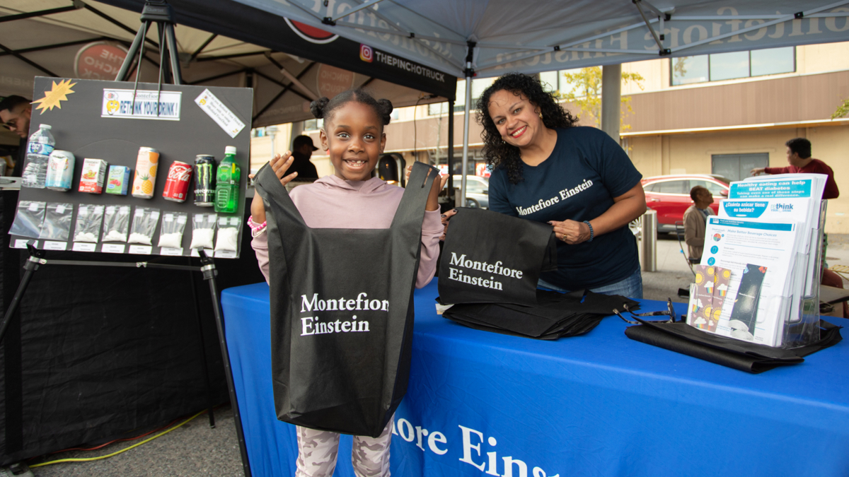 Two participants at the Bronx night market hold up reuseable shopping bags with the Montefiore Einstein logo.