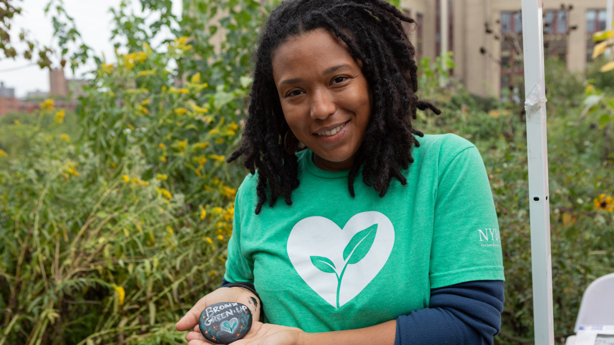A woman smiles and holds up a painted rock that reads "Bronx Green Up".