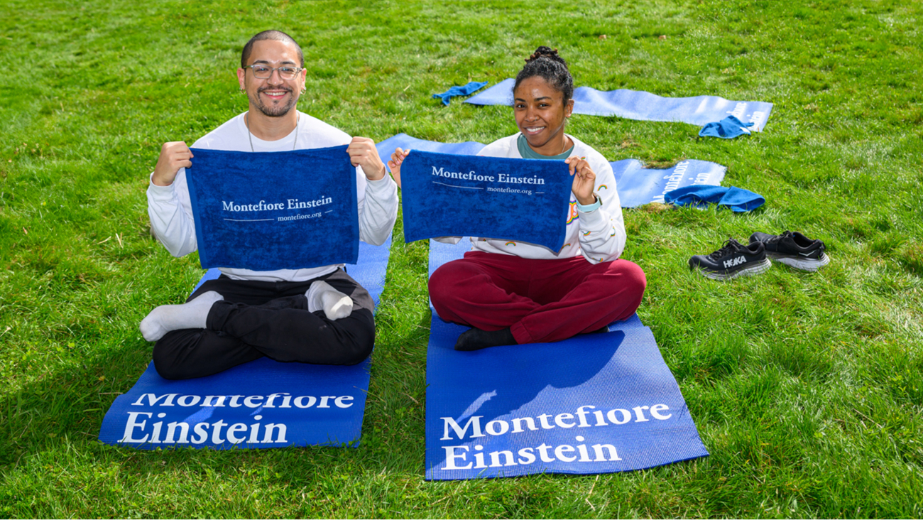 Two smiling participants sit on the ground with Montefiore Einstein towel gifts.