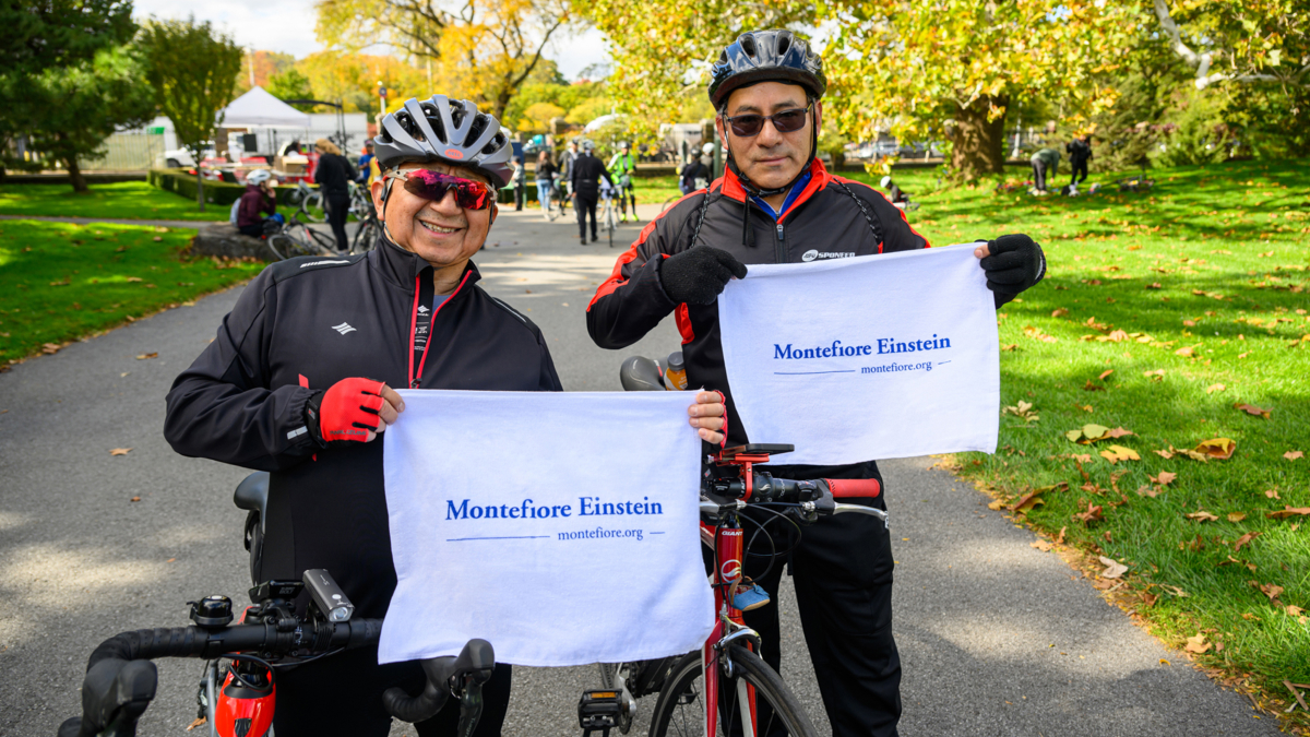 Two road race participants in bike helmets hold up Montefiore Einstein towels.