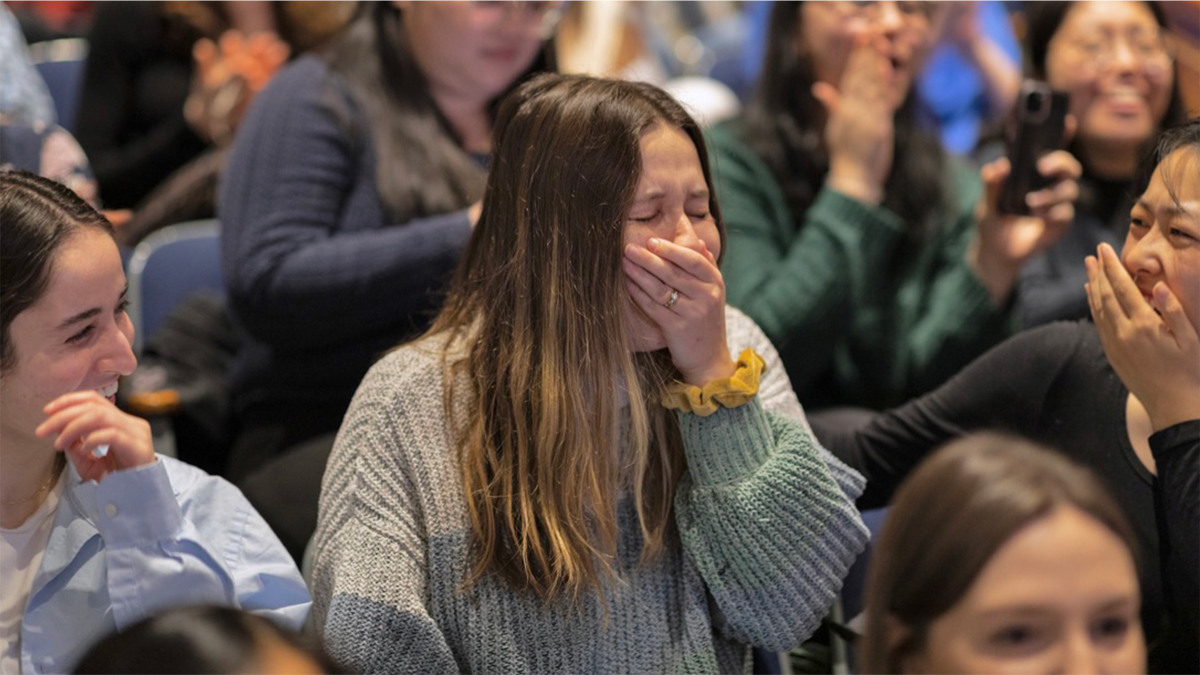 A student covers her mouth with her hand, her eyes closed, as she processes the good news.