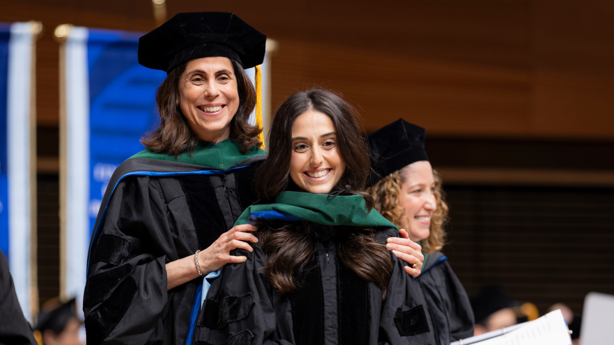 Two smiling students with caps, gowns and green sashes smile toward the camera.