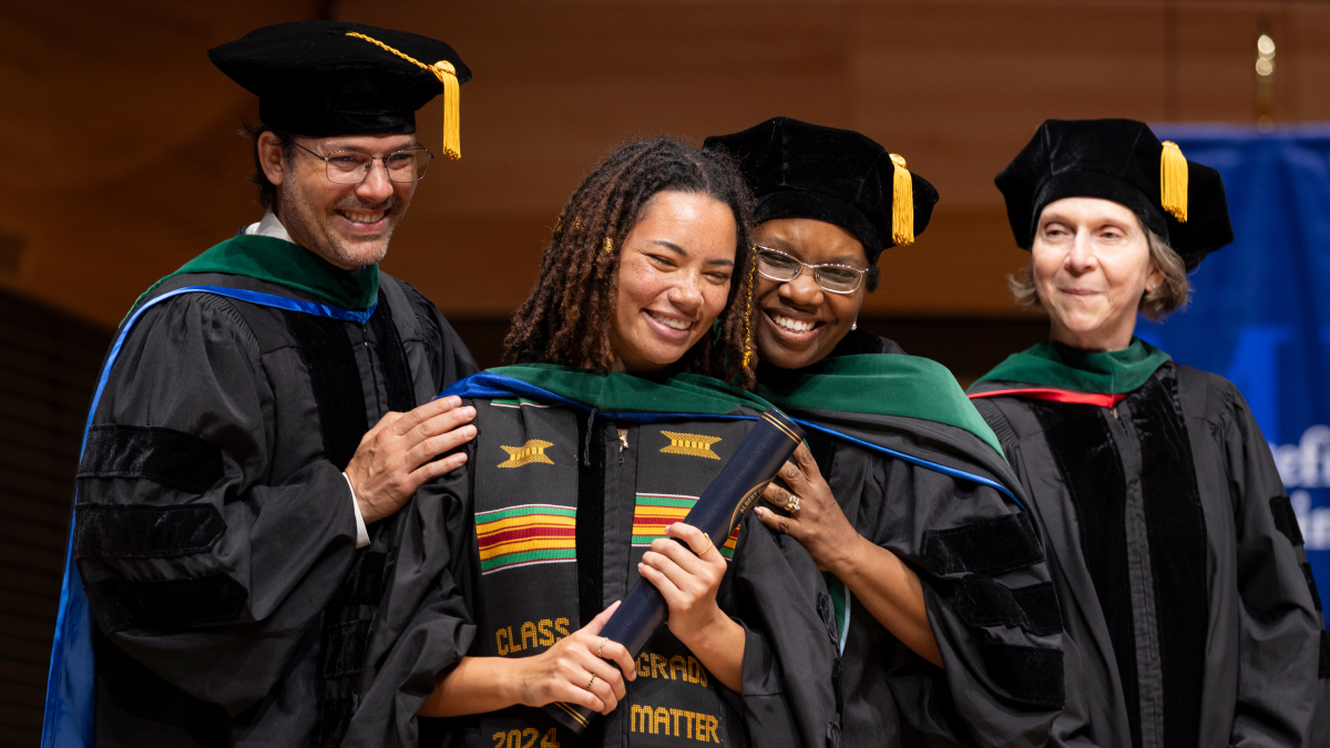 Enthusiastic graduates with green, blue, and multicolored sashes over their gowns smile for a picture.