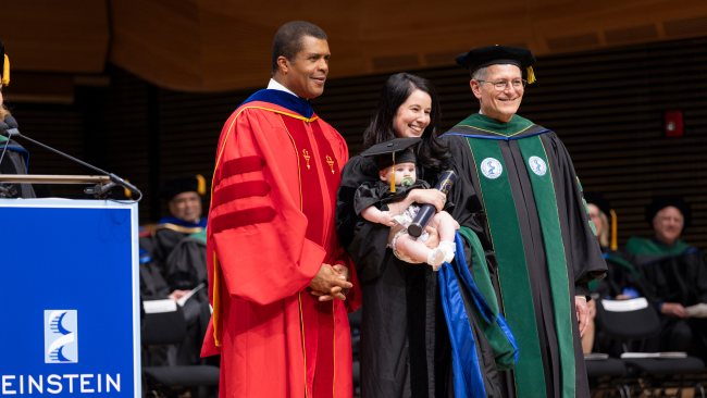 Speakers for commencement stand on the stage, smiling toward the audience.