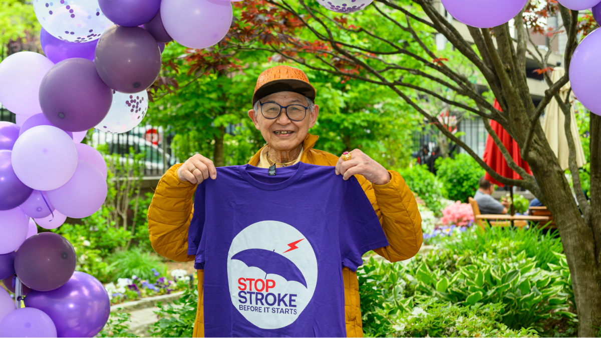 A participant holds up a stroke awareness t-shirt while standing under a lavender and purple balloon arch.