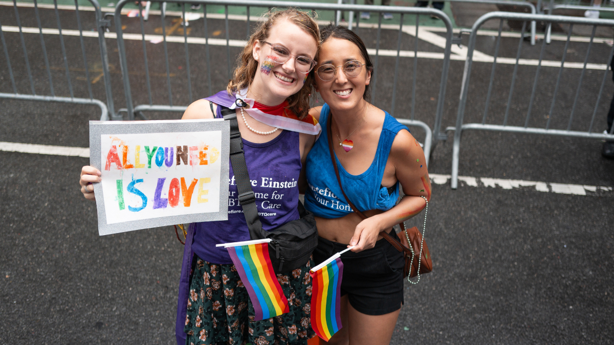 Two parade goers hold rainbow flags and a handmade sign that says "All you need is love." 