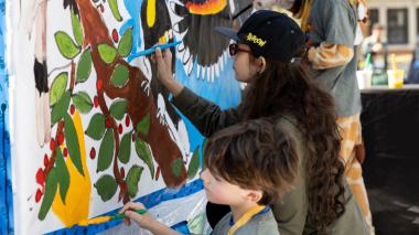 Three children paint a brightly colored large format painting together. The child in the foreground paints a bright yellow fruit, while the next one works on tree leaves.  In the back, the spread feathers of a bird can be seen on a background of sky blue.