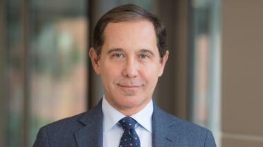 A white man with short brown hair, dressed in a blue suit jacket, light blue shirt, and blue tie looks at the camera with a slight smile.