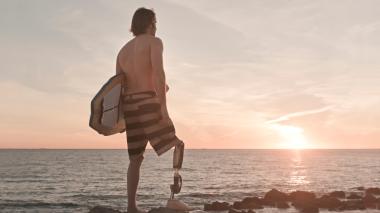 A surfer with a right prosthetic leg watches the sunset on a rocky shore.