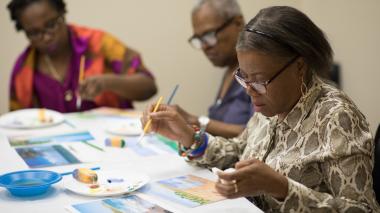 Three participants seated at a table work on landscape paintings.