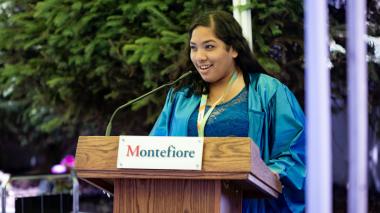 A Project SEARCH graduate in a blue graduation robe speaks at a podium with the Montefiore logo on the front. 