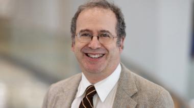 Dr. Ronald C. Samuels, MD, MPH. He smiles toward the camera, wearing a white shirt, maroon tie,  tan jacket and glasses.