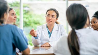 Doctors in white coats and scrubs gather around a table discussing a topic.