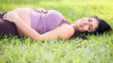 A pregnant woman in a lavender top lies in a shady spot of green grass.  She smiles toward the camera with her hands on her belly.