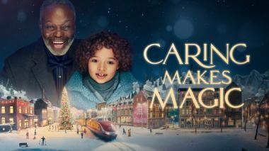 A smiling man and a child with a face full of wonder look down upon a holiday village scene model. The words 'Caring Makes Magic' are to the right in gold.