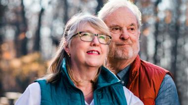 An older man with white hair stands, smiling, behind his wife who looks toward the sky smiling. They're dressed for the fall outdoors, and behind them are a stand of golden-leaved trees. 